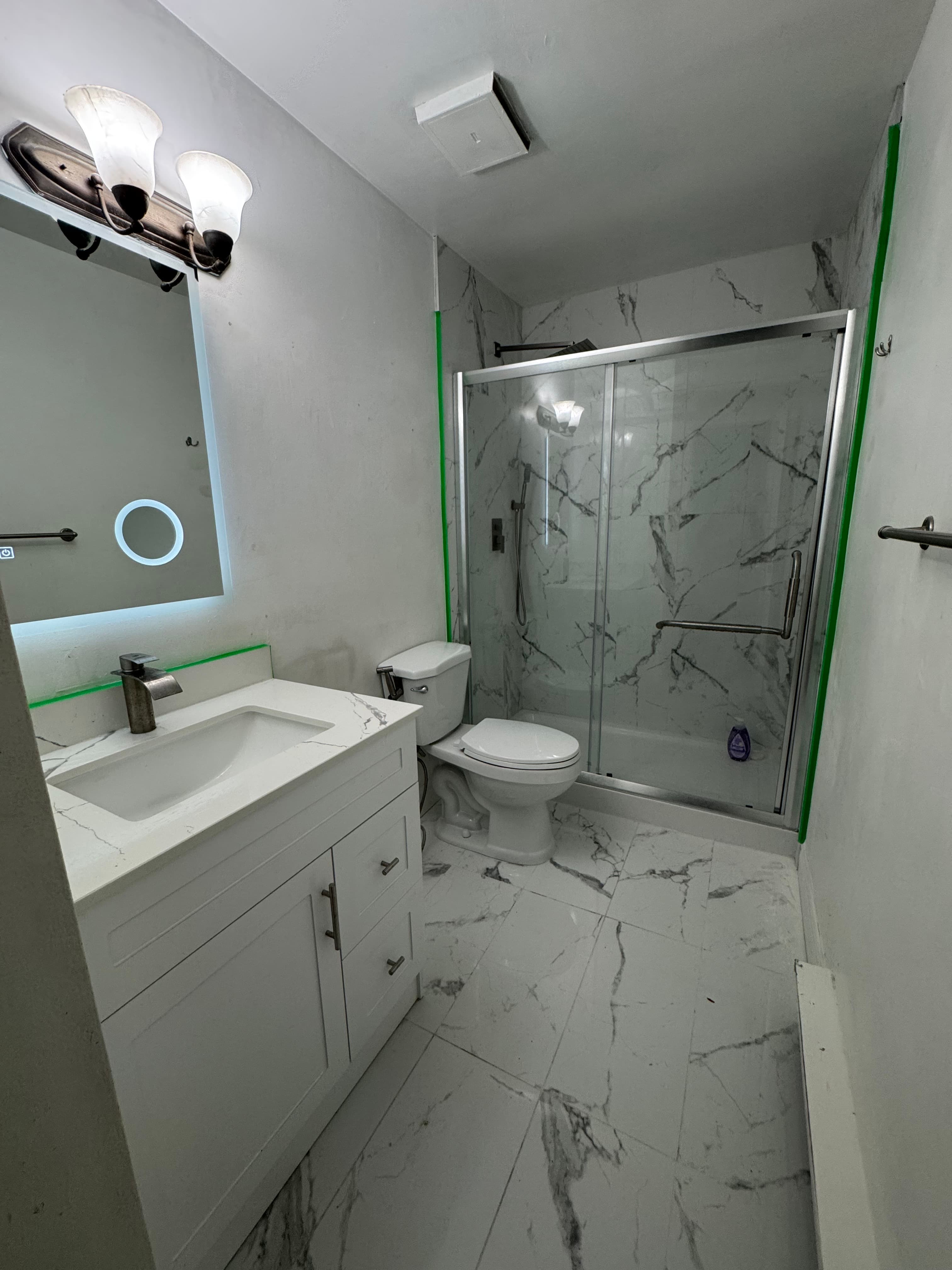 Bathroom during finishing work with a white vanity, illuminated mirror, marble-look shower, and glass doors