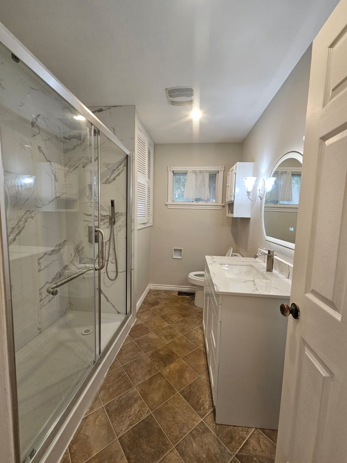 Renovated bathroom with a glass shower, white vanity, illuminated mirror, and warm floor tiles
