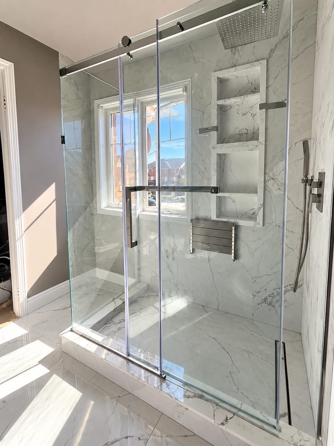 Glass shower enclosure with marble-look tile, built-in shelves, a shower seat, and a rainfall showerhead